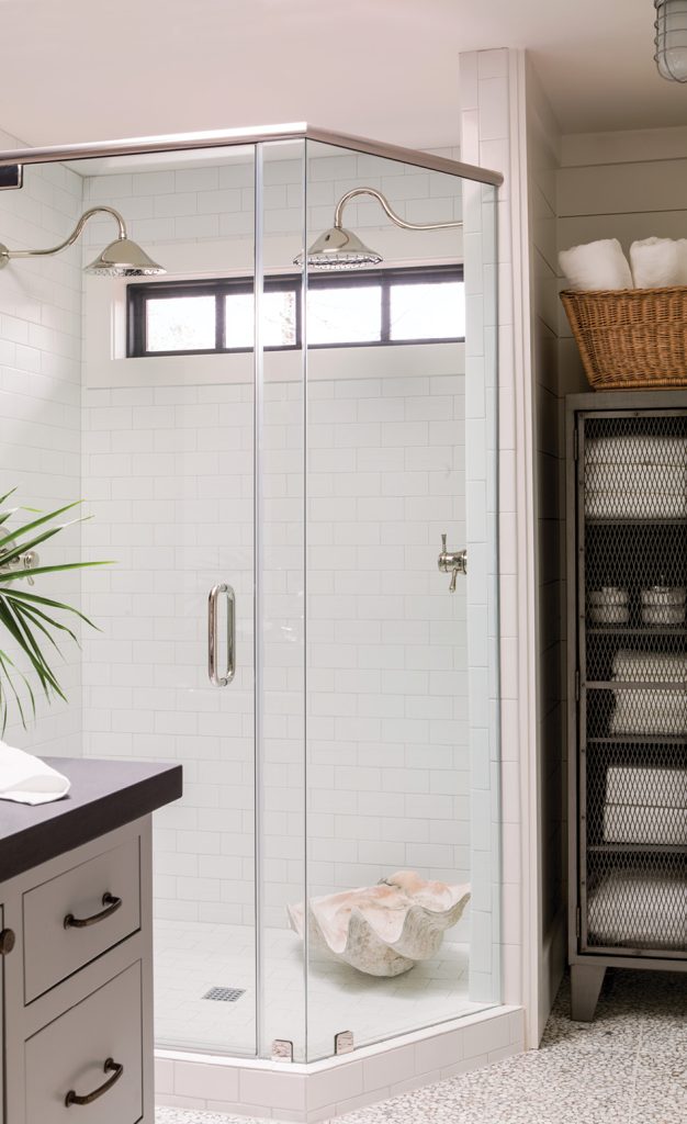 “Symmetry reigns in our family’s lake house bathroom complete with double rain shower heads in polished nickel and a shower flanked by metal cabinets filled with bath supplies. I wanted us to feel as if we’re in a completely different setting when we visit the lake, so I brought in colors and elements that call to mind the water and shore, such as the shiplap walls and the modern gray pebble resin floors. We used an island with a two-sided mirror and sinks in lieu of the traditional vanity because it allowed for better flow.”