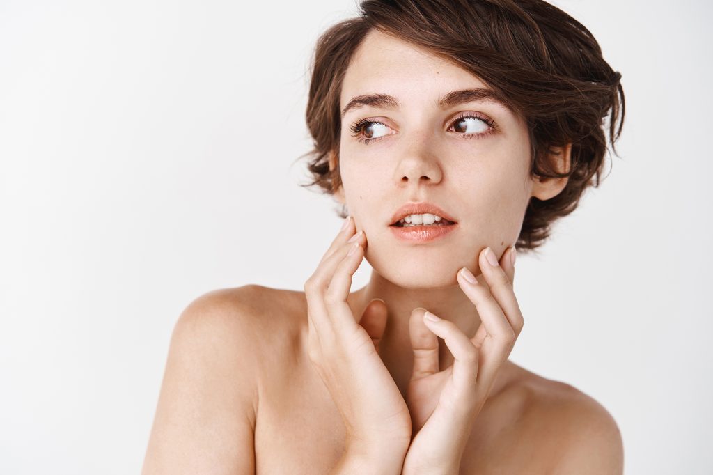 Close up of tender young woman with naked shoulders, gently toughing natural no makeup facial skin, looking away pensive, standing over white background Mastering the No-Makeup Makeup Look Close up of tender young woman with naked shoulders, gently toughing natural no makeup facial skin, looking away pensive, standing over white background