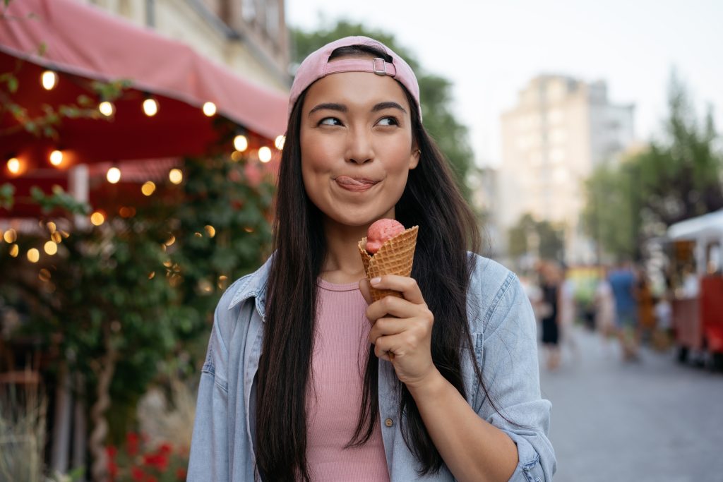Portrait of beautiful asian woman eating ice cream on the street Emotional hipster wearing casual clothing holding tasty summer dessert looking away outdoors Food festival