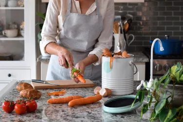 Woman peeling carrots in kitchen and using composter for scraps countertop composting Turn Kitchen Food Scraps into Soil Nutrients in Hours By Annette Brooks